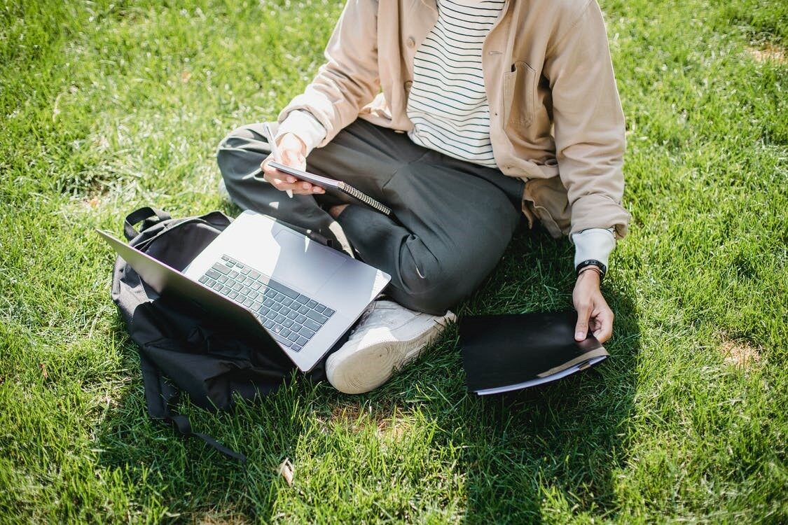 Person sitting on grass with laptop open and holding a notebook and folder